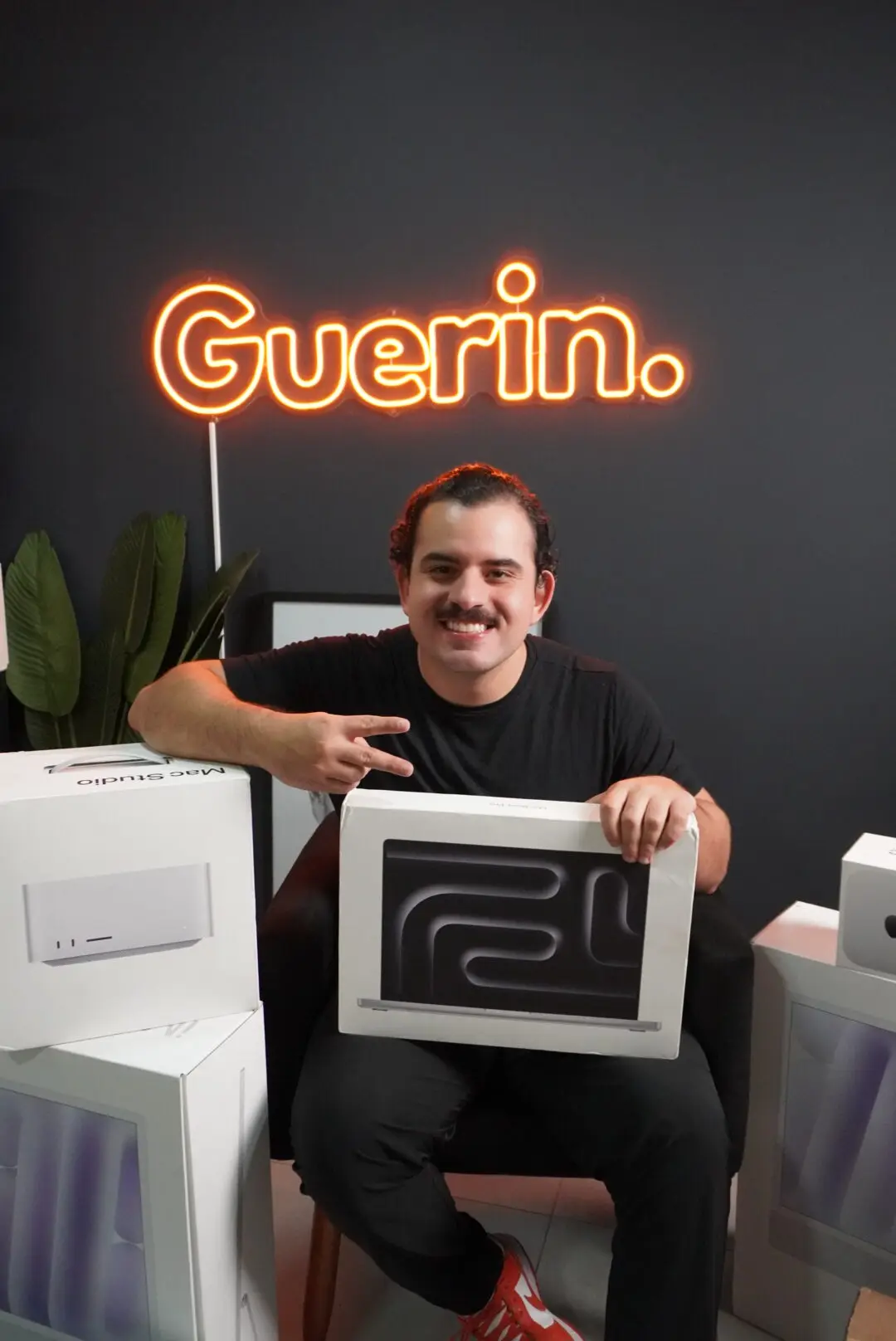 Kevin, fundador da Guerin, sentado em frente ao letreiro neon laranja "Guerin." segurando um MacBook Pro