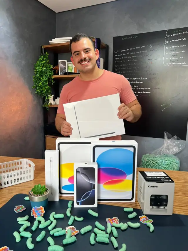 Kevin sorrindo segurando uma caixa de MacBook, com iPad, iPhone, Apple Pencil e lente Canon sobre a mesa e um quadro de encomendas ao fundo